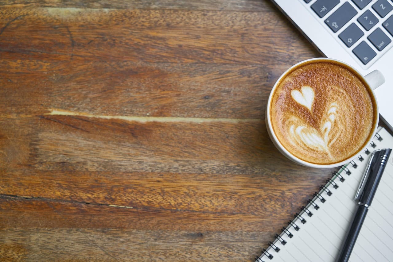 A latte with heart latte art on a wooden desk beside a notebook